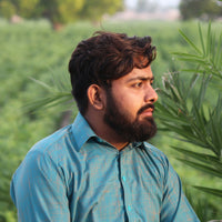 Man in a teal traditional outfit sitting outdoors with greenery in the background