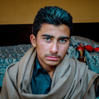 Man wearing a beige shawl over a blue shirt in a room with patterned cushions and a dark headboard.